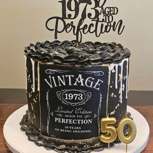 May include: Black and gold cake with a vintage-style label that reads "Vintage 1973 Limited Edition Aged to Perfection 50 Years of Being Awesome". The cake is decorated with black frosting and gold accents. A black and gold topper reads "Since 1973 Aged to Perfection". A gold candle shaped like the number 50 is on top of the cake.