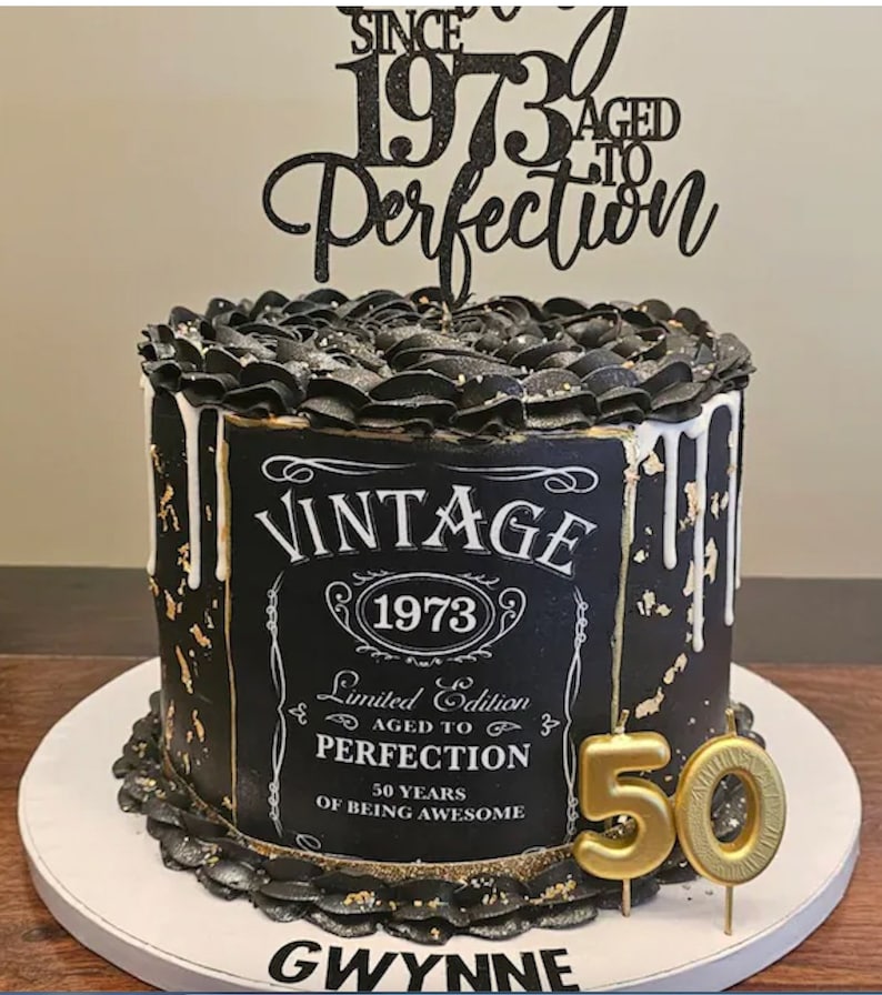 May include: Black and gold cake with a vintage-style label that reads "Vintage 1973 Limited Edition Aged to Perfection 50 Years of Being Awesome". The cake is decorated with black frosting and gold accents. A black and gold topper reads "Since 1973 Aged to Perfection". A gold candle shaped like the number 50 is on top of the cake.