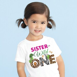 May include: White t-shirt with the text "SISTER of the Wild ONE" in pink and green. The word "ONE" features animal print patterns. The shirt has a jungle theme with a lion, giraffe, and zebra.