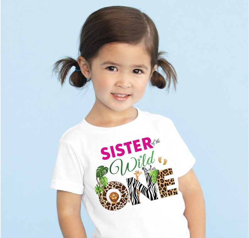 May include: White t-shirt with the text "SISTER of the Wild ONE" in pink and green. The word "ONE" features animal print patterns. The shirt has a jungle theme with a lion, giraffe, and zebra.