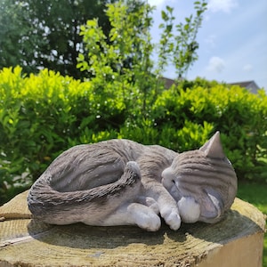 May include: A grey and white sleeping cat figurine. The cat is curled up in a sleeping position with its head resting on its paws. The figurine is made of a smooth, realistic material.