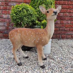May include: A detailed, brown and white spotted deer statue. The deer has a slender neck and is standing on a bed of pebbles. The statue is in front of a brick wall and green shrubbery.