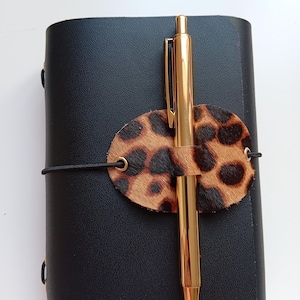 Puede incluir: Un diario de cuero negro con un bolígrafo dorado asegurado por un soporte de cuero con estampado de leopardo. El diario se cierra con una banda elástica negra. El bolígrafo es dorado y tiene un diseño elegante.