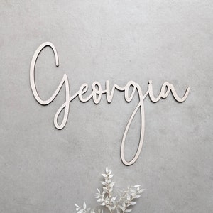 A personalised wooden name sign in a handwritten calligraphy design, in this example for Georgia. The name is cut from birch plywood in our Ash font.  The wooden name is presented on a neutral background with dried flower accents.