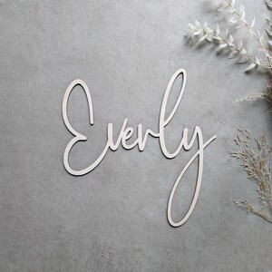 A personalised wooden name sign in a handwritten calligraphy design, in this example for Everly. The name is cut from birch plywood in our Ash font.  The wooden name sign is presented on a neutral background with dried flower accents.