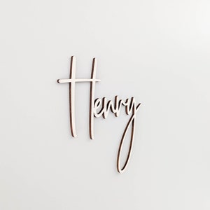 A personalised wooden name sign in a handwritten calligraphy design, in this example for Henry. The name is cut from birch plywood in our Ash font.  The wooden name is presented on a neutral background.