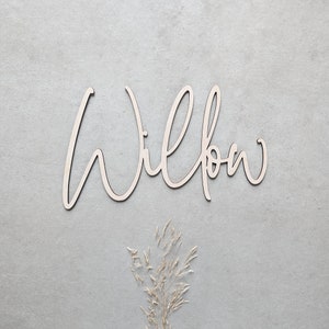 A personalised wooden name sign in a handwritten calligraphy design, in this example for Willow. The name is cut from birch plywood in our Ash font.  The wooden name is presented on a neutral background with dried flower accents.