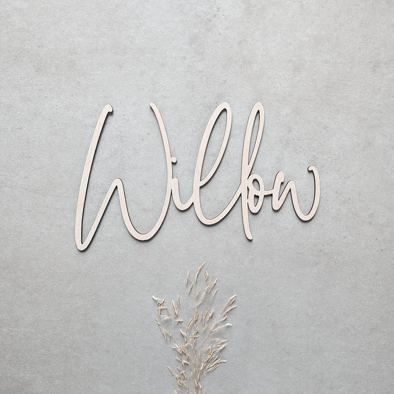 A personalised wooden name sign in a handwritten calligraphy design, in this example for Willow. The name is cut from birch plywood in our Ash font.  The wooden name is presented on a neutral background with dried flower accents.