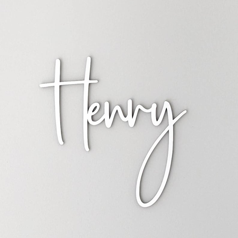 A personalised wooden name sign in a handwritten calligraphy design, in this example for Henry. The name is cut from birch plywood in our Ash font.  The wooden name is shown painted in white mounted on a grey wall.