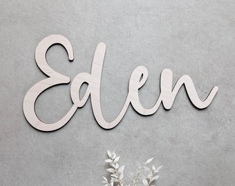 May include: Wooden sign spelling "Eden" in a cursive font. The sign is made of light-colored wood with a dark outline, and is placed on a gray background. A sprig of white decorative leaves is at the bottom.