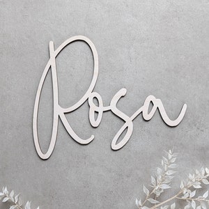 A personalised wooden name sign in a handwritten calligraphy design, in this example for Rosa. The name is cut from birch plywood in our Ash font.  The wooden name is presented on a neutral background with dried flower accents.