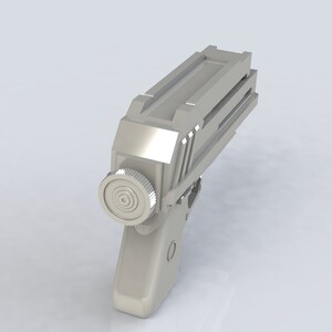 K&ouml;nnte beinhalten: Eine silberfarbene, futuristisch aussehende Pistole mit einem zylindrischen Bauteil an der Seite und einer geriffelten Oberseite. Die Waffe ist vor einem schlichten wei&szlig;en Hintergrund platziert.