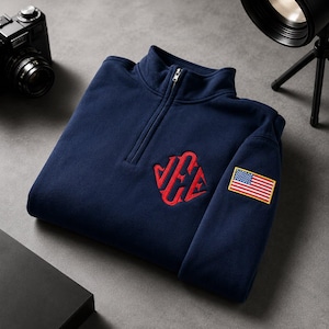 Peut inclure: Un pull bleu marine à fermeture éclair quart, orné d'un monogramme rouge brodé sur la poitrine et d'un petit écusson représentant le drapeau américain sur la manche. Le vêtement possède un col montant avec une fermeture éclair argentée.