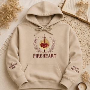 May include: A beige hooded sweatshirt featuring an embroidered design on the chest of a sword piercing a flaming heart surrounded by a laurel wreath. The text FIREHEART is below the design. The sleeves feature the text TO WHATEVER END and YOU DO NOT YIELD.