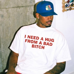 May include: A white short-sleeved t-shirt featuring the red text I NEED A HUG FROM A BAD BITCH printed on the chest. The garment is paired with a solid blue baseball cap that displays a colourful graphic logo on the front panel above the brim.