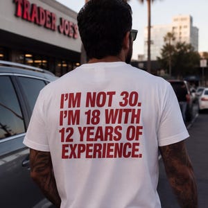 Può includere: Una t-shirt bianca a maniche corte con un testo rosso in grassetto sul retro che recita: I'M NOT 30. I'M 18 WITH 12 YEARS OF EXPERIENCE. La maglia presenta un girocollo e una vestibilità standard, con il messaggio stampato chiaramente sulla schiena.