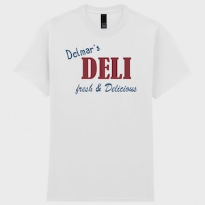 May include: A white short-sleeve t-shirt featuring the text Delmar's DELI fresh & Delicious printed on the front. The word DELI is in large red capital letters, while Delmar's and fresh & Delicious are in a smaller blue script font.