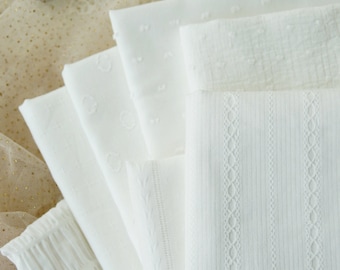 May include: A collection of folded white fabric swatches featuring various textures and patterns, including raised dots, vertical stripes, and intricate woven lace-like details, arranged in a fan shape alongside a piece of sheer gold glitter fabric.