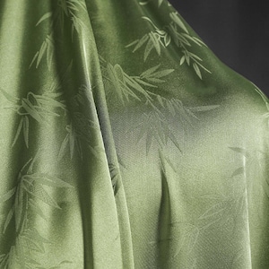 Pode incluir: Close-up de um tecido verde-oliva fluido com um brilho sutil. O tecido apresenta um padrão repetido de folhas e caules de bambu. As dobras e sombras criam profundidade e textura, destacando a queda do tecido.