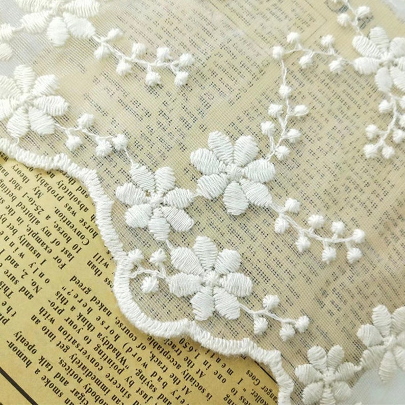 May include: White lace trim with a delicate floral pattern. The lace is on a white background and has a scalloped edge.
