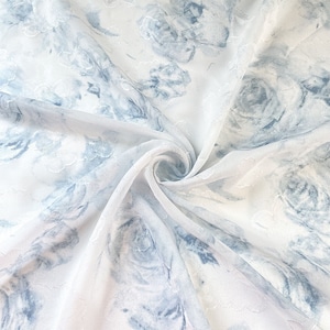 May include: White sheer fabric with a blue floral pattern. The fabric is gathered and twisted, showing the texture and drape of the material.