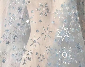 May include: A sheer, light blue tulle fabric featuring a repeating pattern of glittery blue snowflakes, white outlined stars, and small white circular dots scattered across the surface, creating a textured and layered visual effect.
