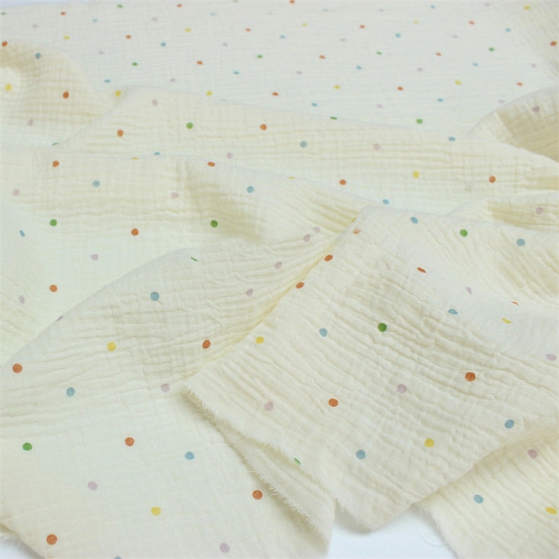 May include: Off-white fabric with a crinkled texture, adorned with small, multi-coloured polka dots. The dots are in shades of red, green, blue, and yellow. The fabric is folded, displaying its soft, flowing quality. Suitable for various craft projects.