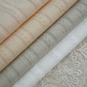 May include: Four different types of fabric, including a white, a light grey, a peach, and a beige fabric with a textured pattern. The fabrics are stacked on top of each other, with the white fabric on the bottom and the beige fabric on the top.