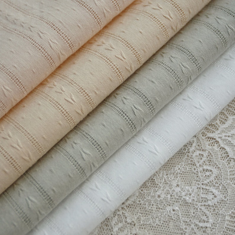 May include: Four different types of fabric, including a white, a light grey, a peach, and a beige fabric with a textured pattern. The fabrics are stacked on top of each other, with the white fabric on the bottom and the beige fabric on the top.