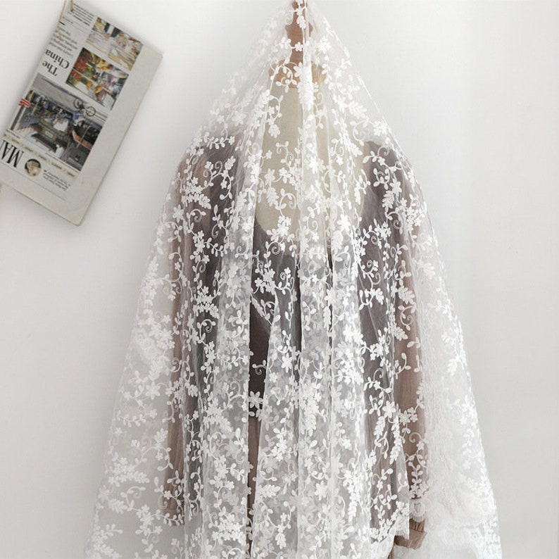 May include: A sheer white lace fabric featuring an intricate floral embroidery pattern with delicate vines and small blossoms. The lightweight material is draped over a mannequin, showcasing the detailed needlework and semi-transparent texture.