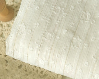 May include: Close-up of a folded piece of off-white fabric with a textured surface. The fabric has a vertical striped pattern with raised floral-like details. A wooden spool is visible in the background, suggesting a sewing or crafting theme.