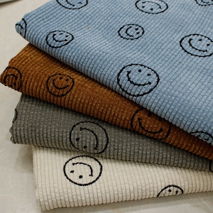 Könnte beinhalten: Ein Stapel Cordstoff in Blau-, Braun-, Grau- und Cremetönen. Jedes Stück ist mit einem sich wiederholenden Muster aus schwarzen Smiley-Gesichtern bedruckt. Der Stoff hat eine gerippte Textur.
