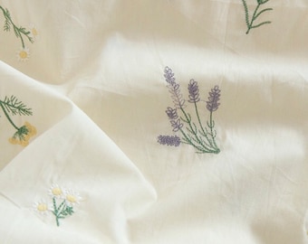 May include: A cream-colored fabric featuring scattered embroidered floral motifs. The design includes small clusters of yellow flowers, white daisies with yellow centers, and purple lavender sprigs, all connected by thin green stems and leaves.