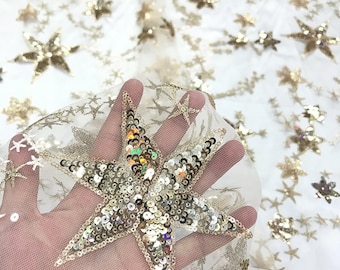 May include: A sheer white tulle fabric featuring an all-over pattern of embroidered gold stars. The stars vary in size and are embellished with reflective gold sequins that catch the light, creating a shimmering effect across the lightweight mesh material.