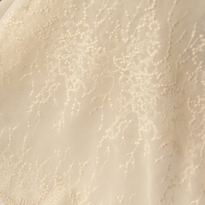 Peut inclure: Gros plan sur un tissu d&eacute;licat de couleur cr&egrave;me avec un motif floral subtil. Le tissu est transparent, presque translucide, et le motif floral est cr&eacute;&eacute; avec de petits points en relief.
