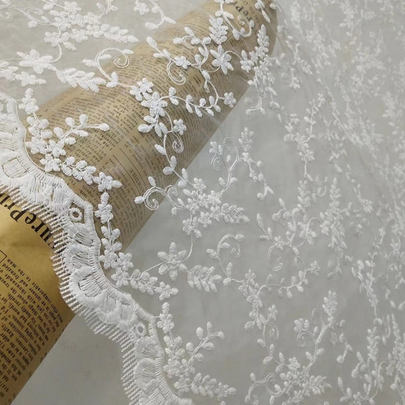 May include: A sheer white fabric featuring intricate floral and vine embroidery with a scalloped edge. The lace is draped over a cylindrical roll of paper printed with vintage-style black text, showcasing the delicate needlework and pattern.