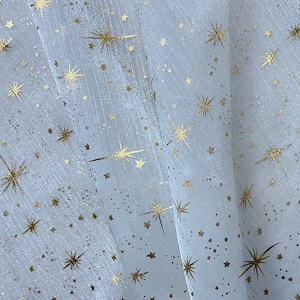 Könnte beinhalten: Nahaufnahme eines transparenten, hellfarbenen Stoffes mit einem sich wiederholenden Muster aus goldenen Sternen und kleinen Punkten. Die Sterne variieren in der Größe und haben mehrere Spitzen, wodurch ein himmlisches Design entsteht. Der Stoff ist leicht durchscheinend.