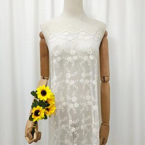 May include: White embroidered fabric with a delicate floral pattern. The fabric is sheer and has a scalloped edge.