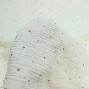 May include: Cream-coloured muslin fabric with a crinkled texture and a pattern of colourful polka dots. The dots are in shades of orange, green, blue, yellow, and purple. The fabric is soft and lightweight, ideal for baby blankets or clothing.