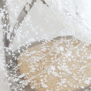 May include: A sheer white fabric featuring intricate white floral and vine embroidery. The delicate material displays a repeating pattern of small flowers and leafy stems, creating a lightweight, semi-transparent textile with a detailed texture.