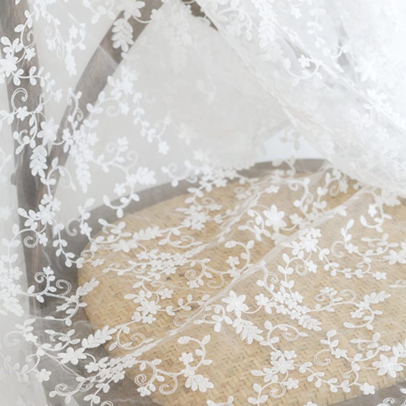 May include: A sheer white fabric featuring intricate white floral and vine embroidery. The delicate material displays a repeating pattern of small flowers and leafy stems, creating a lightweight, semi-transparent textile with a detailed texture.