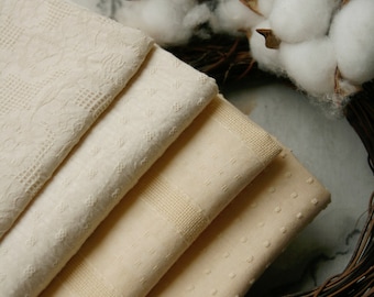 May include: A stack of four folded fabric swatches in varying shades of cream and off-white. Each piece of fabric features a distinct textured pattern, including floral lace, small embroidered dots, and woven grid designs on the textile surfaces.