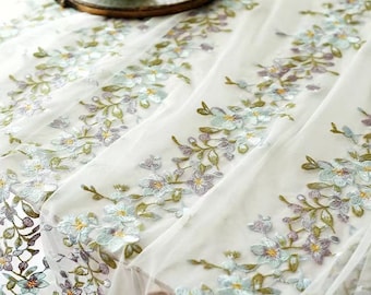 May include: A sheer white table runner featuring intricate embroidery of floral vines in shades of light blue, lavender, and olive green. The delicate fabric displays a repeating pattern of blossoms and leaves arranged in vertical bands across the surface.