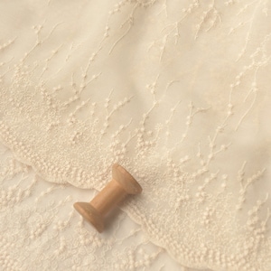 Peut inclure: Gros plan sur une dentelle d&eacute;licate de couleur blanc cass&eacute; avec un motif floral. Le tissu est doux et transparent, avec un l&eacute;ger brillant. Une bobine de fil en bois repose sur le tissu.