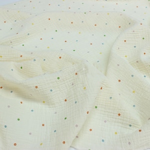 May include: Off-white fabric featuring a polka dot design. The dots are in a variety of colours, such as orange, green, blue, and yellow. The fabric has a textured, crinkled look, possibly muslin or gauze. The fabric is draped and folded.