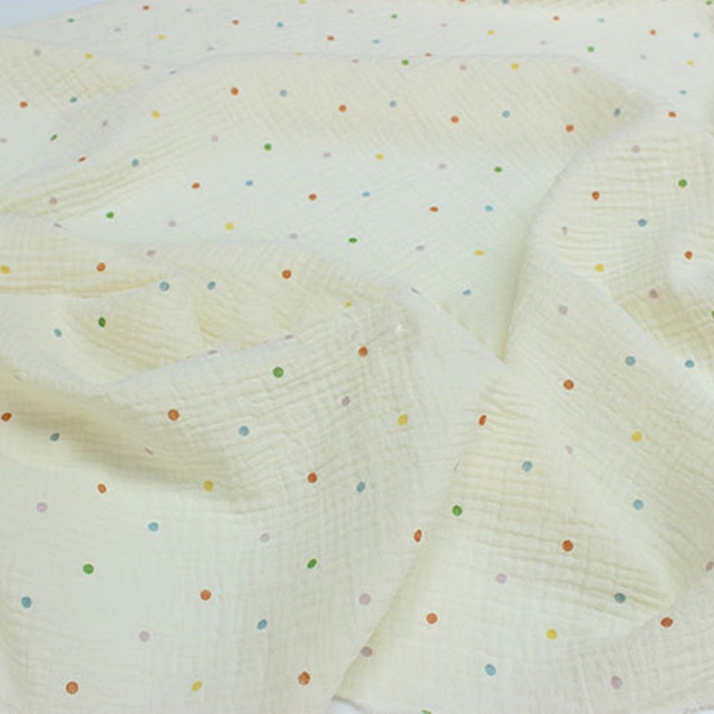 May include: Off-white fabric featuring a polka dot design. The dots are in a variety of colours, such as orange, green, blue, and yellow. The fabric has a textured, crinkled look, possibly muslin or gauze. The fabric is draped and folded.