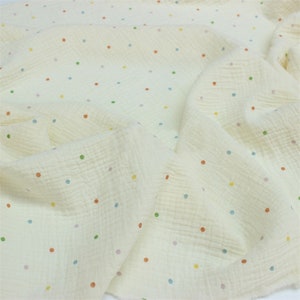 May include: Cream-coloured muslin fabric with a crinkled texture, scattered with small, colourful polka dots in shades of red, green, blue, and yellow. The fabric is draped, showing its soft, flowing nature.