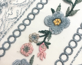 May include: A sheer white mesh fabric featuring intricate embroidery of light blue and pink flowers with green leaves, accented by vertical rows of circular blue trim and delicate, swirling light blue thread patterns throughout the textile.