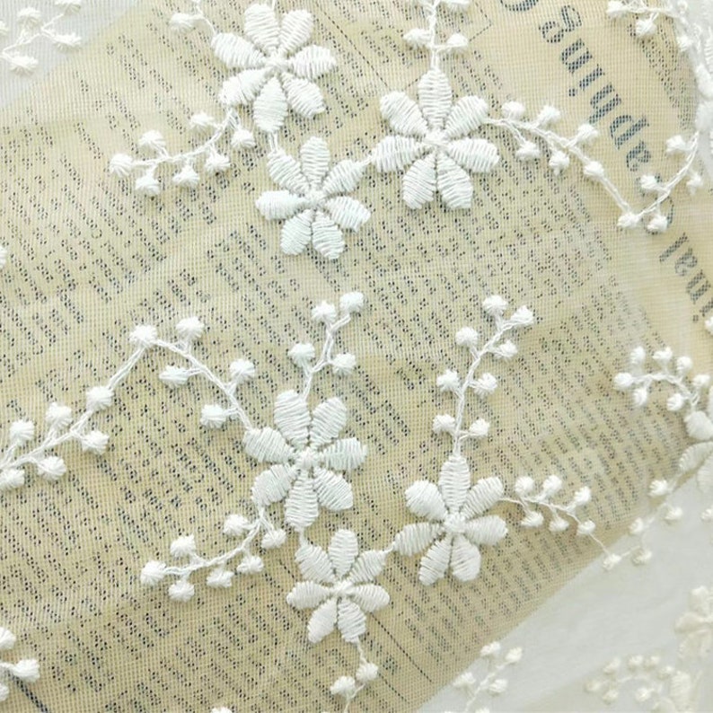 May include: White floral lace fabric with a delicate pattern of small flowers and leaves. The lace is sheer and has a soft, romantic feel.