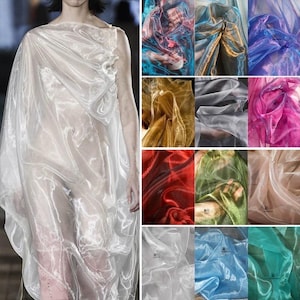 May include: A sheer, iridescent organza fabric shown in a variety of colors including white, blue, gold, red, green, pink, and teal. The material features a translucent, shimmering quality with a crisp texture that drapes into soft folds.